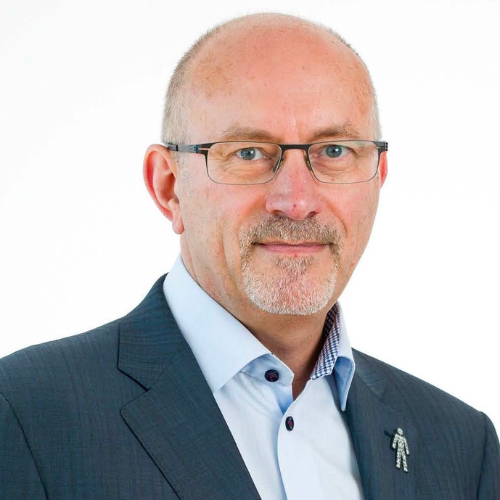 A white man with a light white beard and glasses, smiling. He wears an unbuttoned shirt, suit jacket and a Prostate Cancer UK badge.