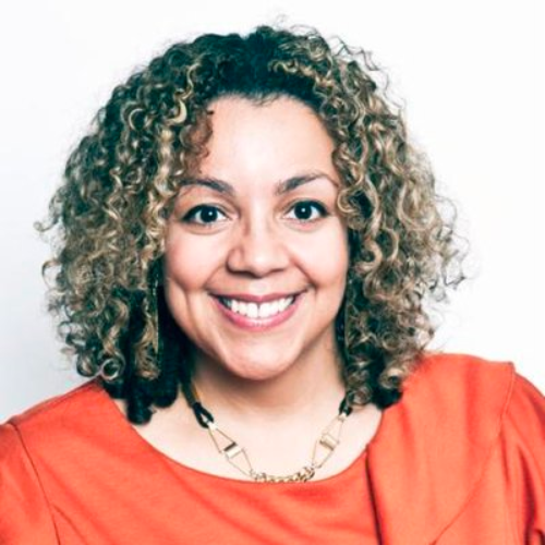 A biracial Black woman with highlighted, curly hair is smiling at the camera. She wears an orange to, and a beaded necklace.