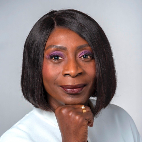 A Black woman in her late forties/early fifties smiles at the camera, resting her chin on her hand. She has a straightened, shiny bob hairstyle, and purple eye shadow and red lipstick. She looks very glamorous.